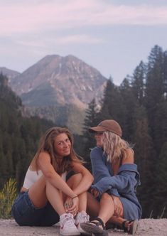 two women sitting on top of a mountain next to each other with trees in the background