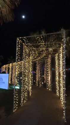 a walkway covered in lights at night with palm trees and moon in the sky behind it