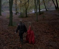 two people walking in the woods with leaves on the ground and one person wearing a red cloak