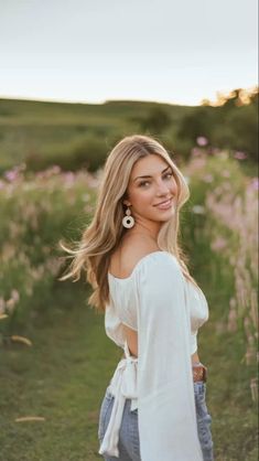 a beautiful blonde woman standing in a field with flowers on the side and her hair blowing in the wind