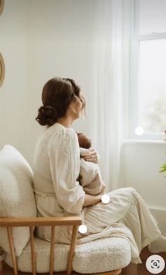 a woman sitting on a chair holding a baby in her arms and looking out the window