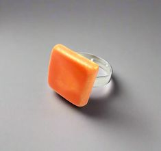 an orange square shaped ring sitting on top of a gray surface with a silver band