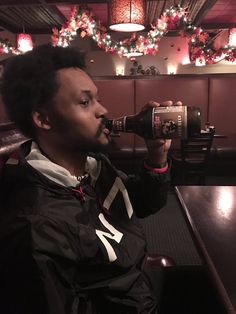 a man sitting at a table drinking from a beer bottle in front of christmas lights