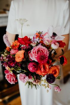 a woman holding a bouquet of flowers in her hands and wearing a white dress with red, pink, orange and purple flowers