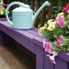 a blue watering pot sitting on top of a purple wooden bench next to pink and yellow flowers