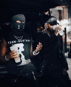 two men in black outfits and masks drinking from cups while sitting on the back of a car