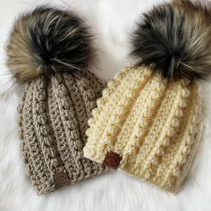 two crocheted hats with faux fur pom - poms are laying on a white blanket