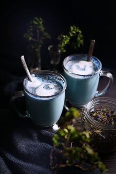 two mugs filled with blue liquid sitting on top of a table next to plants