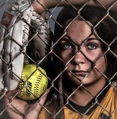 a woman holding a baseball in her right hand behind a chain link fence and looking at the camera