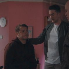 three men standing in a room with pink walls and one is pointing at the wall