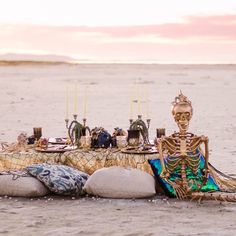 a skeleton sitting on top of a sandy beach next to a table with candles and other items