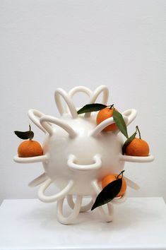 a white vase with oranges in it sitting on top of a table next to a wall