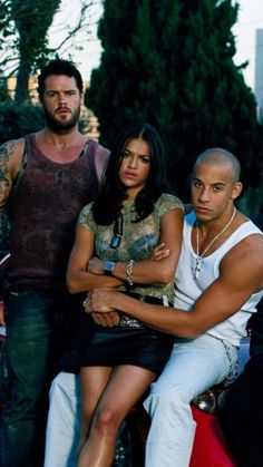 two men and a woman sitting on a motorcycle with their arms around each other's shoulders