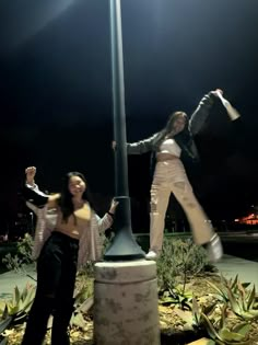 two women standing next to each other near a street light pole at night with their arms in the air