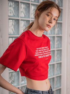 a woman wearing a red crop top with white writing on the front and side, standing in front of a glass block wall