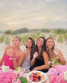 four women sitting at a table with food and drinks in front of them on the beach