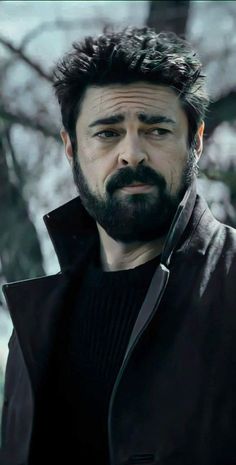 a man with a beard wearing a black jacket and looking at the camera while standing in front of a tree