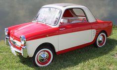 an old red and white car is parked in the grass near some water on a cloudy day