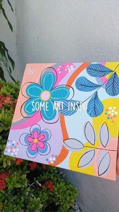 a person holding up a colorful flowered painting in front of a potted plant