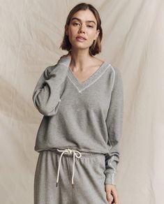 A person wearing The V-Neck Sweatshirt in Varsity Grey by THE GREAT. FALL 24 KNITS and drawstring pants stands against a beige background. V-neck Sweatshirt With Ribbed Cuffs For Loungewear, Cozy V-neck Sweatshirt For Loungewear, Cozy V-neck Sweatshirt With Ribbed Cuffs, Emily And Meritt, Wing Shoes, Red Wing Shoes, College Sweatshirt, Deep V Neck, Raglan Sleeve