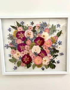 a white frame with flowers in it on the wall next to a vase filled with blue and pink flowers