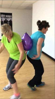 two women are doing exercises with balloons in an office building, one is holding a purple ball and the other has a green shirt