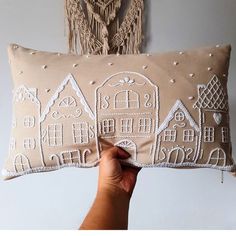 a hand holding a pillow with embroidered houses on it and fringes hanging from the wall