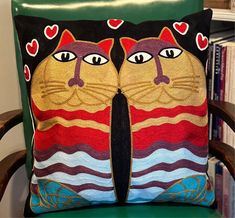 two decorative pillows with cats on them sitting on a green chair in front of a bookshelf