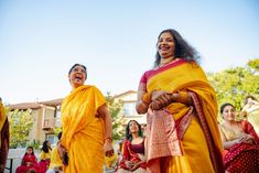 two women in yellow and red sari are laughing at each other while others look on