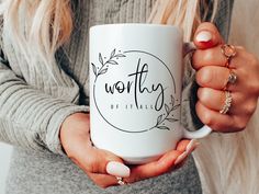 a woman holding a white coffee mug with the word worthy on it's side