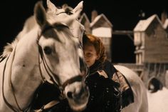 a woman standing next to two white horses in the snow with houses in the background