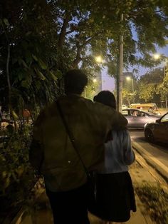 two people walking down the sidewalk at night with cars parked on the street behind them