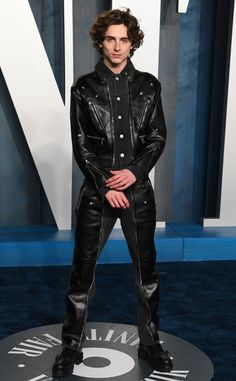 a man in black leather suit standing on blue carpet with his hands behind his back