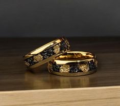 two gold wedding bands with black diamonds on them sitting on a wooden table next to each other