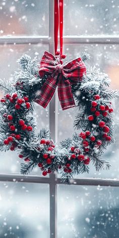 a christmas wreath hanging on a window sill in front of a snowy scene with evergreens and red berries