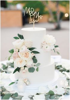 a wedding cake with white flowers and greenery on top is decorated with a mr and mrs sign