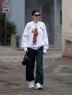 a woman walking down the street wearing a white sweatshirt and green pants