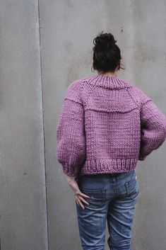 a woman standing in front of a wall with her back turned to the camera, wearing a purple sweater and jeans