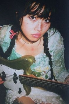 a young woman holding a green and white electric bass guitar in her right hand while looking at the camera