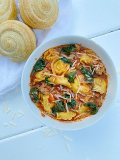 a white bowl filled with pasta and spinach on top of a table next to rolls