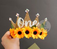 a hand holding up a sunflower crown with the word one on it and two flowers