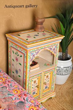 an ornate painted cabinet next to a potted plant on a bed with pink and yellow bedspread