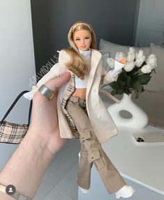 a hand holding a barbie doll in front of a table with white flowers and a vase