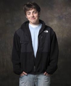 a young man standing in front of a black background with his hands in his pockets