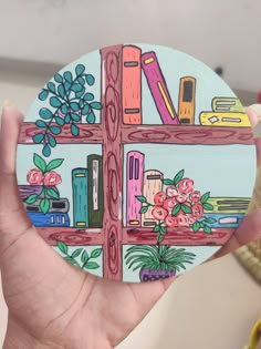 a hand holding up a painted coaster with books and flowers on the shelf behind it