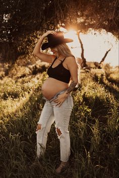 a pregnant woman standing in the grass with her hands on her head and wearing a hat