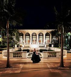 a woman standing in front of a building at night with palm trees and lights on