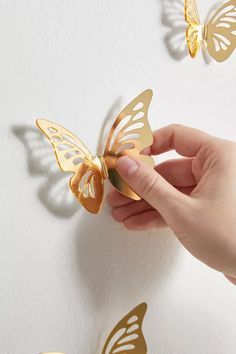 a hand holding a golden butterfly on top of a white wall with gold butterflies flying around it