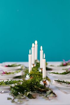 a table topped with white plates covered in greenery and lit candles next to each other