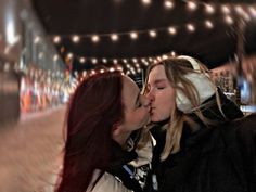 two women kissing each other in front of string lights on a city street at night